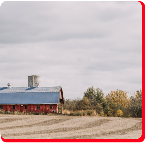 A weathered red barn with a blue metal roof sits on an open field surrounded by autumn trees near Toronto, showcasing a candidate for historic barn restoration.