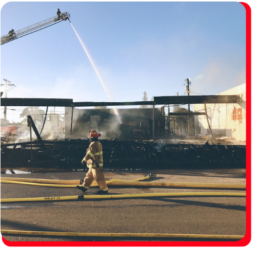 Firefighter walking past a severely burned commercial building as fire crews use a ladder hose to extinguish remaining flames—representing emergency commercial fire cleanup and restoration efforts Firefighter walking past a severely burned commercial building as fire crews use a ladder hose to extinguish remaining flames—representing emergency commercial fire cleanup and restoration efforts