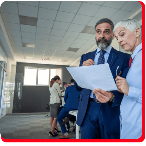 Two professionally dressed people discuss a document in an office specializing in commercial restoration, with two others working at a desk in the background. The modern workspace, featuring large windows and fluorescent lights, reflects a professional environment dedicated to services like water damage restoration, fire damage restoration, commercial water damage restoration, and commercial fire restoration.