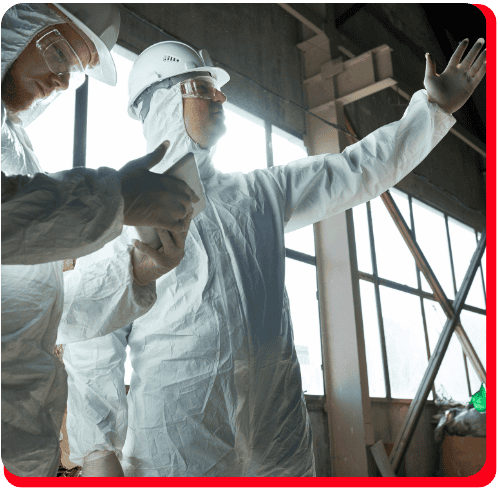 Two workers in white protective suits, gloves, hard hats, and safety glasses stand indoors at a commercial fire restoration site; one points forward while the other uses a tablet to assess fire damage restoration progress. Large windows and industrial equipment emphasize the commercial environment, highlighting ongoing commercial water damage restoration and related cleanup efforts.
