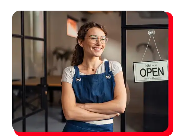 A smiling woman in glasses and a blue apron stands with folded arms beside a glass door displaying an OPEN sign, looking confident inside a modern shop specializing in commercial restoration, including fire damage restoration, water damage restoration, commercial fire restoration, and commercial water damage restoration services.