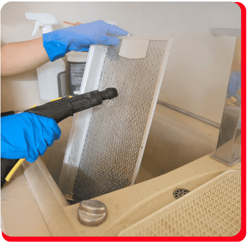 A person wearing blue gloves uses a handheld steam cleaner to clean a metal mesh filter over a sink, an essential process in commercial restoration including fire damage restoration and water damage restoration, especially as part of commercial fire restoration and commercial water damage restoration.