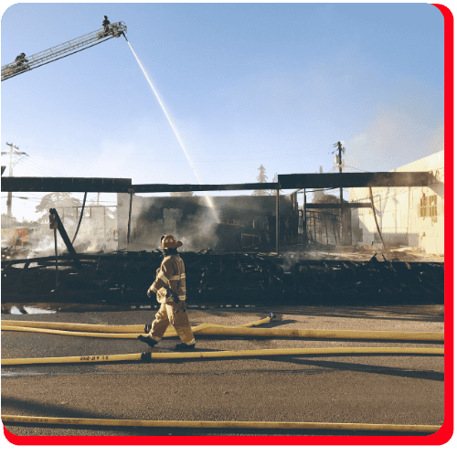 Firefighter walking past a severely burned commercial building as fire crews use a ladder hose to extinguish remaining flames—representing emergency commercial fire cleanup and restoration efforts