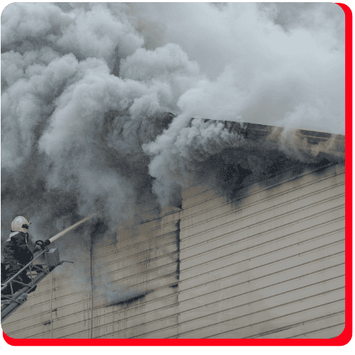 A firefighter on a ladder sprays water at a burning building with thick gray smoke pouring from the roof, demonstrating the immediate need for commercial restoration services such as commercial fire restoration, fire damage restoration, and commercial water damage restoration.