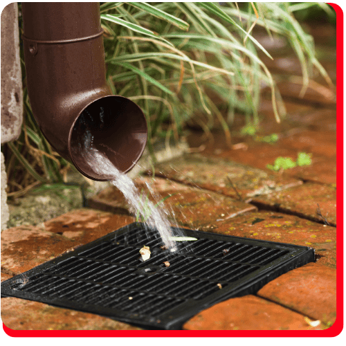Water flows from a brown downspout onto a black drain grate, surrounded by wet red bricks and green plants in the background—highlighting essential infrastructure for commercial water damage restoration, water damage restoration, and broader commercial restoration needs.