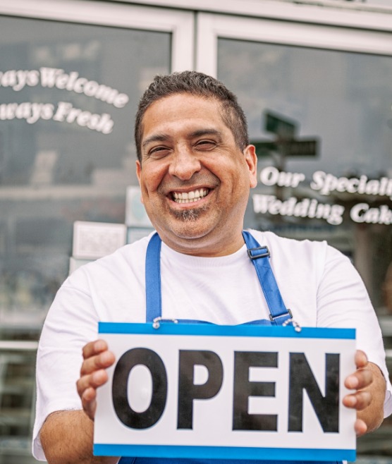 Smiling man in a blue apron stands outside a shop, holding an OPEN sign, representing commercial restoration services. Behind him, glass doors and welcome signs suggest expertise in commercial fire restoration and commercial water damage restoration, highlighting fire damage restoration and water damage restoration solutions for businesses.