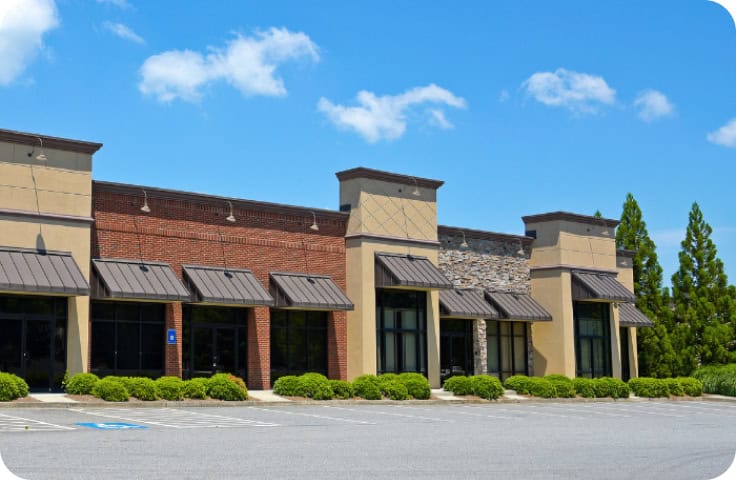 A row of vacant storefronts in a modern strip mall with large windows, brick and stone facades, and metal awnings sits beside a mostly empty parking lot beneath a blue sky—an optimal location for commercial restoration services including fire damage restoration, water damage restoration, commercial fire restoration, and commercial water damage restoration.