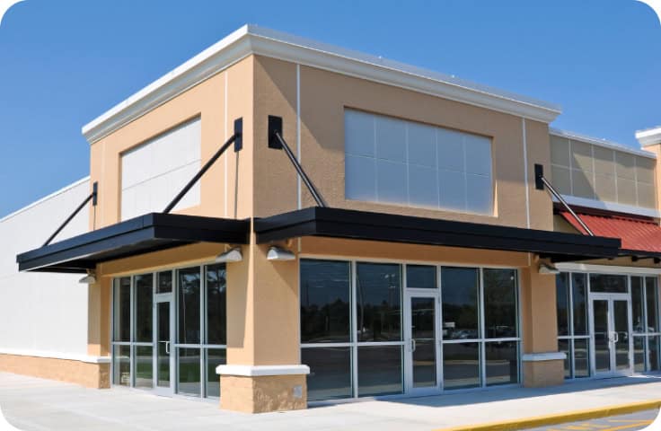 A vacant retail storefront featuring large glass windows and doors, beige walls, and black awnings beneath a clear blue sky—an optimal site for commercial restoration projects such as fire damage restoration, water damage restoration, commercial fire restoration, or commercial water damage restoration.