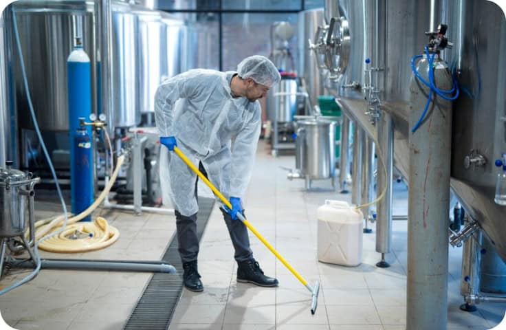 A worker in protective clothing and a hairnet is mopping the floor of an industrial facility as part of commercial water damage restoration. The scene highlights best practices for commercial restoration, fire damage restoration, and water damage restoration in large facilities, with stainless steel tanks and hoses visible in the background to emphasize commercial fire restoration capabilities.