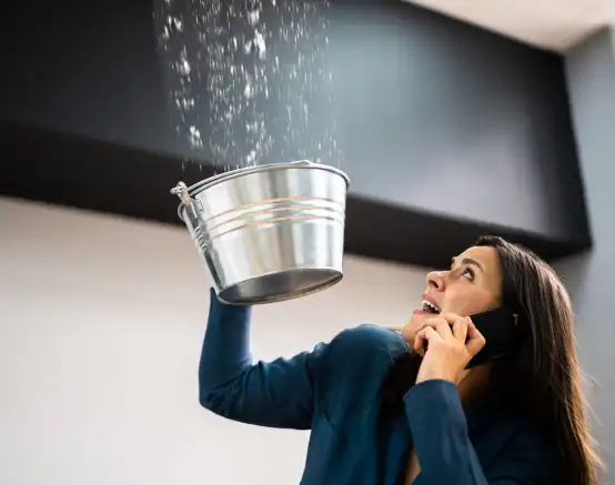 A worried woman holds a metal bucket under a leaking ceiling to catch dripping water while talking on the phone, depicting an urgent situation that highlights the need for commercial restoration services, particularly commercial water damage restoration or fire damage restoration.