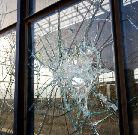 A close-up of a window with a large, circular crack and shattered glass, reflecting light and hinting at commercial restoration needs. The blurred view of a building structure outside suggests damage potentially requiring fire damage restoration, water damage restoration, or specialized commercial fire and water damage restoration services.