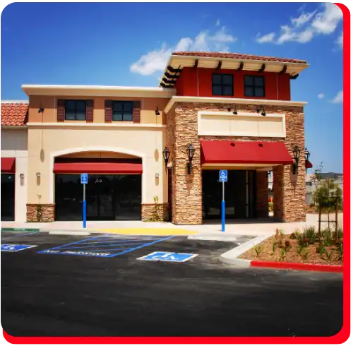 A modern shopping center featuring stone accents, red awnings, and large windows has two wheelchair-accessible parking spaces at the entrance. This location is ideal for companies in need of commercial restoration services such as water damage restoration, fire damage restoration, commercial water damage restoration, or commercial fire restoration. Blue sky with a few clouds is visible above the building.