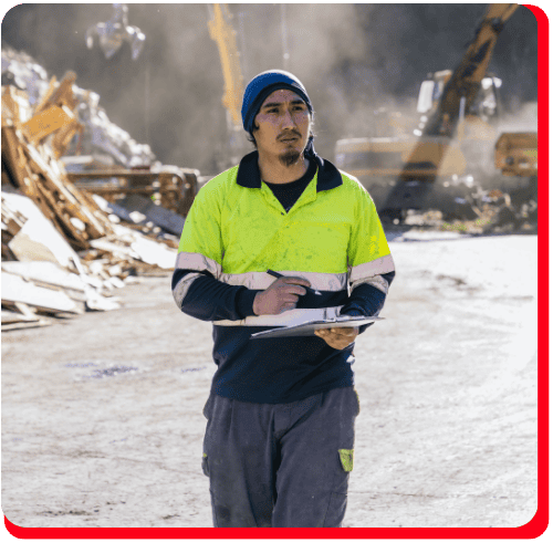 A construction worker in a yellow safety shirt and blue beanie holds a clipboard while surveying a commercial restoration site, with excavators and debris scattered throughout the dusty area, highlighting activities related to fire damage restoration and water damage restoration.