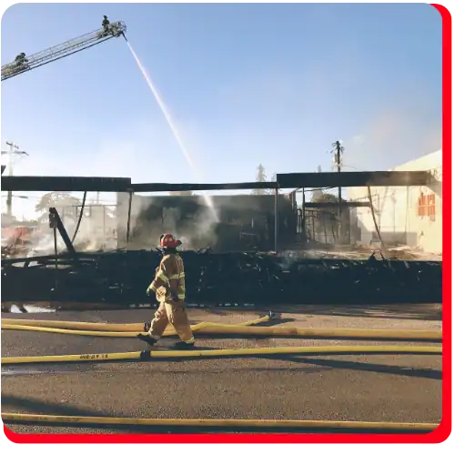 Firefighters perform commercial fire restoration as one walks past a severely charred building and another sprays water from above to aid in fire damage restoration and water damage restoration. Smoke billows from the burnt commercial structure, with yellow hoses laid out on the street, highlighting extensive commercial water damage restoration efforts.