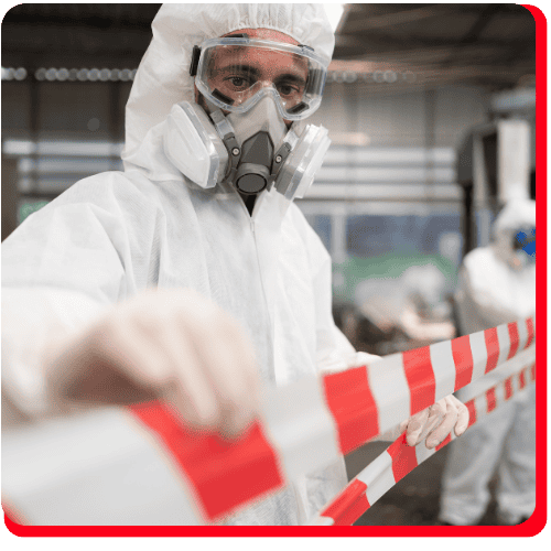 Technician in full protective suit and gas mask holds red-and-white caution tape inside an industrial facility undergoing commercial restoration, with another restoration specialist visible but blurred in the background—environment suggests ongoing fire damage restoration or water damage restoration as part of comprehensive commercial fire or water damage restoration services.