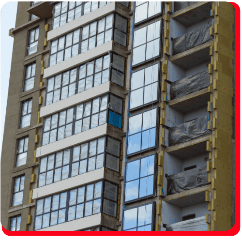 A tall commercial building features numerous glass windows and balconies, with some areas covered by plastic sheets indicating recent commercial water damage restoration or fire damage restoration efforts. Signs of professional commercial restoration services are visible, suggesting both commercial water damage restoration and possible commercial fire restoration. One window stands out in blue among the clear ones. The image is framed with a red border featuring rounded corners.