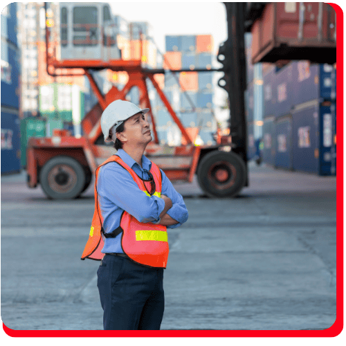 A person in a white hard hat and orange safety vest stands with arms crossed at a shipping yard, surveying stacked containers and machinery commonly found in commercial restoration settings such as fire damage restoration, water damage restoration, commercial fire restoration, and commercial water damage restoration projects.