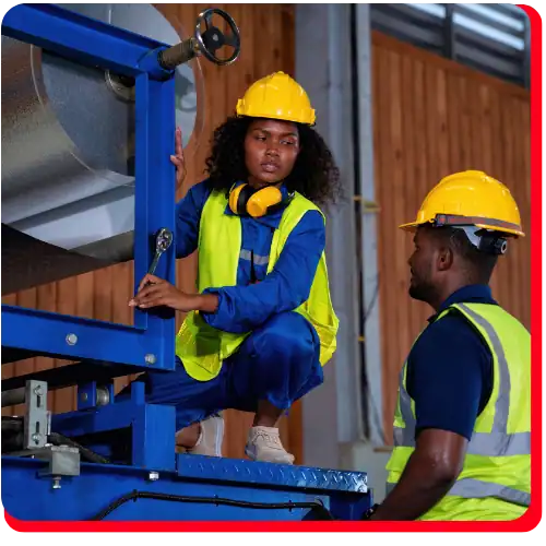 Two workers in yellow hard hats and reflective vests are discussing commercial restoration procedures while repairing machinery in an industrial environment. One worker is crouched on the equipment holding a wrench, possibly performing fire damage restoration or water damage restoration, while the other stands facing them, collaborating on commercial fire restoration or commercial water damage restoration tasks.