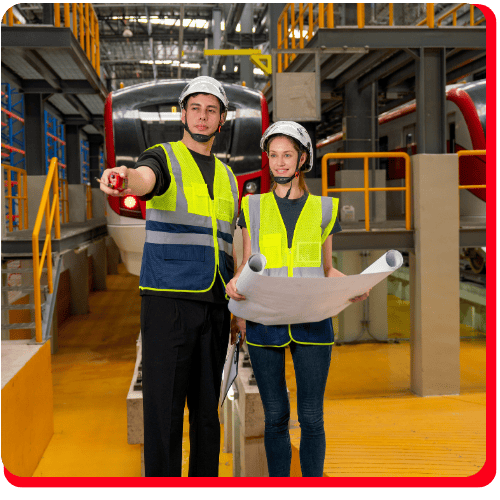 Two engineers in safety vests and helmets stand in front of a train inside an industrial maintenance facility, surrounded by yellow railings and machinery related to commercial restoration. One engineer points ahead while the other holds blueprints for fire damage restoration, with visible elements suggesting commercial fire restoration and commercial water damage restoration work within the environment.