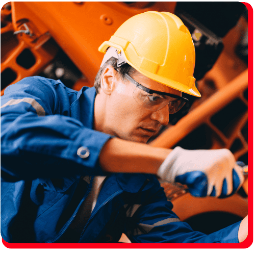 A worker in a yellow hard hat, safety glasses, and blue uniform uses a tool to perform maintenance on machinery as part of a commercial restoration process, focusing on equipment involved in fire damage restoration or water damage restoration; orange machine parts suggest the setting is equipped for commercial fire restoration and commercial water damage restoration services.