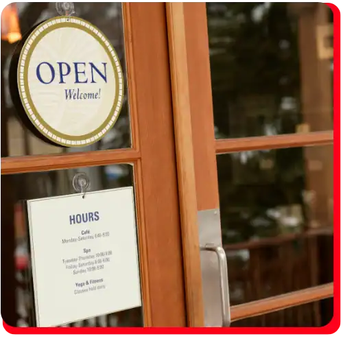 A glass door with an OPEN Welcome! sign and business hours notice is displayed, featuring a metal handle. In the blurred background, an outdoor scene sets the context for a typical business entryway that may need commercial restoration services, such as commercial water damage restoration or commercial fire restoration after incidents requiring fire damage restoration or water damage restoration solutions.
