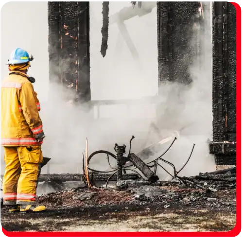 A firefighter in yellow gear stands beside the charred remains of a commercial building, with smoke rising and a burned bicycle among the debris, highlighting the need for fire damage restoration and commercial fire restoration services.