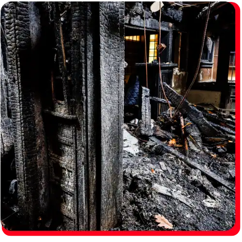A severely fire-damaged room with charred wooden beams, burnt debris, and ashes illustrates the extensive need for commercial fire restoration and fire damage restoration. Warm light glows from a window in the background, contrasting with the destruction and underscoring the importance of professional commercial restoration services for both fire damage restoration and potential water damage restoration following firefighting efforts.