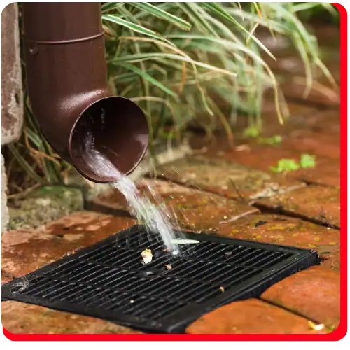 Water pours from a brown gutter downspout onto a black grate in red brick pavement, framed by green plants in the background—a visual reminder of how prompt commercial water damage restoration, and broader commercial restoration services, are crucial to preventing extensive damage. This scene highlights the need for immediate response with water damage restoration after an emergency and underscores commercial fire restoration and fire damage restoration as essential elements of comprehensive property protection.