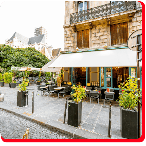 A quaint Parisian café with outdoor seating, potted plants, and a striped awning sits on a cobblestone street next to a historic building—recently revitalized with expert commercial fire restoration and commercial restoration services. The restored property features charming shuttered windows and iron balconies, reflecting recovery from previous fire damage restoration; nearby evidence of commercial water damage restoration highlights comprehensive preservation.