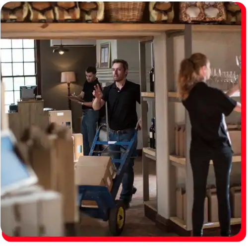 In a storage room typical of commercial restoration environments, three people collaborate: one man uses a hand truck to move boxes—an essential step in fire damage restoration and water damage restoration; another man documents inventory on a clipboard, while a woman organizes glassware on shelves. The organized space, filled with cardboard boxes and shelving, reflects the processes involved in commercial fire restoration and commercial water damage restoration.
