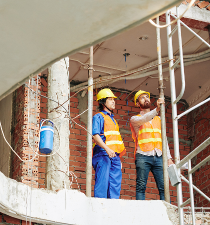 Two construction workers in yellow hard hats and orange safety vests stand on a commercial restoration site, surrounded by scaffolding and exposed brick walls, closely observing ongoing fire damage restoration or water damage restoration work as part of a commercial fire restoration or commercial water damage restoration project.