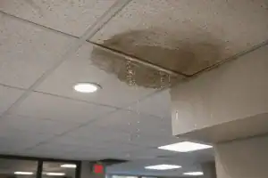 Water leaks from a stained ceiling tile, forming a puddle on the floor below, illustrating the need for commercial water damage restoration. Surrounding tiles and ceiling lights are visible, emphasizing indoor water damage commonly addressed in commercial restoration and commercial fire restoration services.